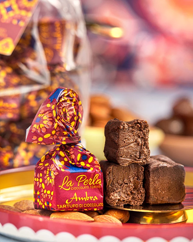 Ambra Trüffelpraline im Ornament-Papier mit drei geöffneten Pralinen auf goldenem Teller